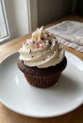 A single chocolate cupcake with white frosting and red and white peppermint pieces on top.