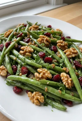 A white serving bowl filled with roasted green beans, cranberries, and walnuts.